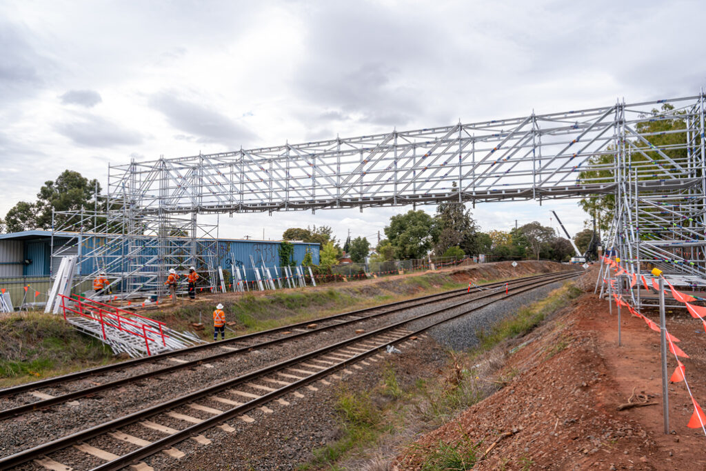 A steel scaffold bridge stretches high over a railway line in an urban area.