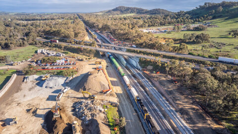 Aerial view of rail tracks passing under a road bridge with surrounding earthworks and construction equipment.