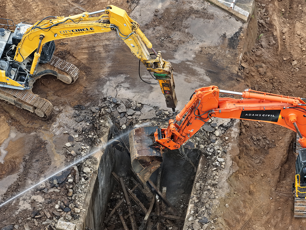 Excavators breaking up a concrete bridge structure inside a trench.
