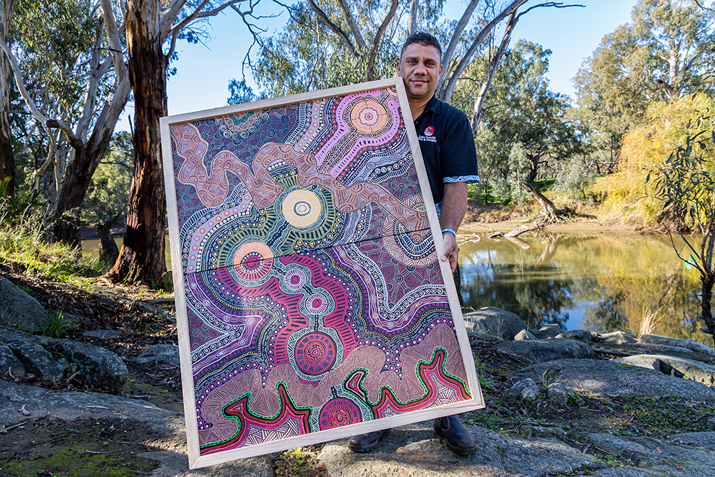 Person holding a framed artwork beside a creek surrounded by trees.