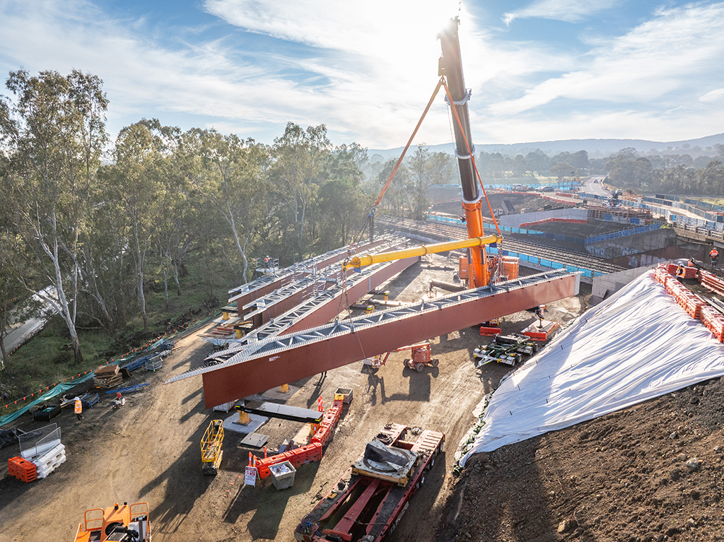 Crane lifting a steel bridge girder from a transport trailer at a construction site.