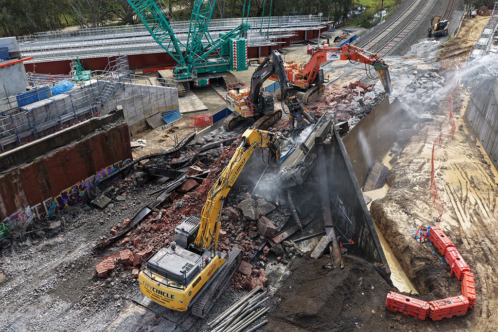 Excavators demolishing a bridge structure beside railway tracks with cranes and construction equipment nearby.