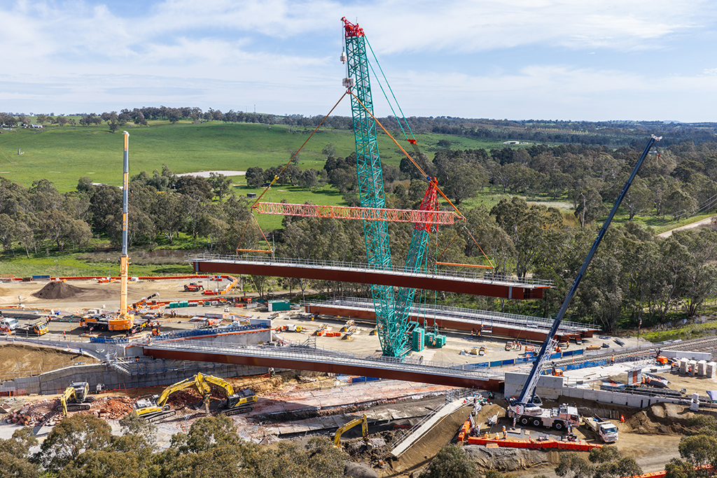 Large crane lifting a steel bridge girder into place above a construction site.