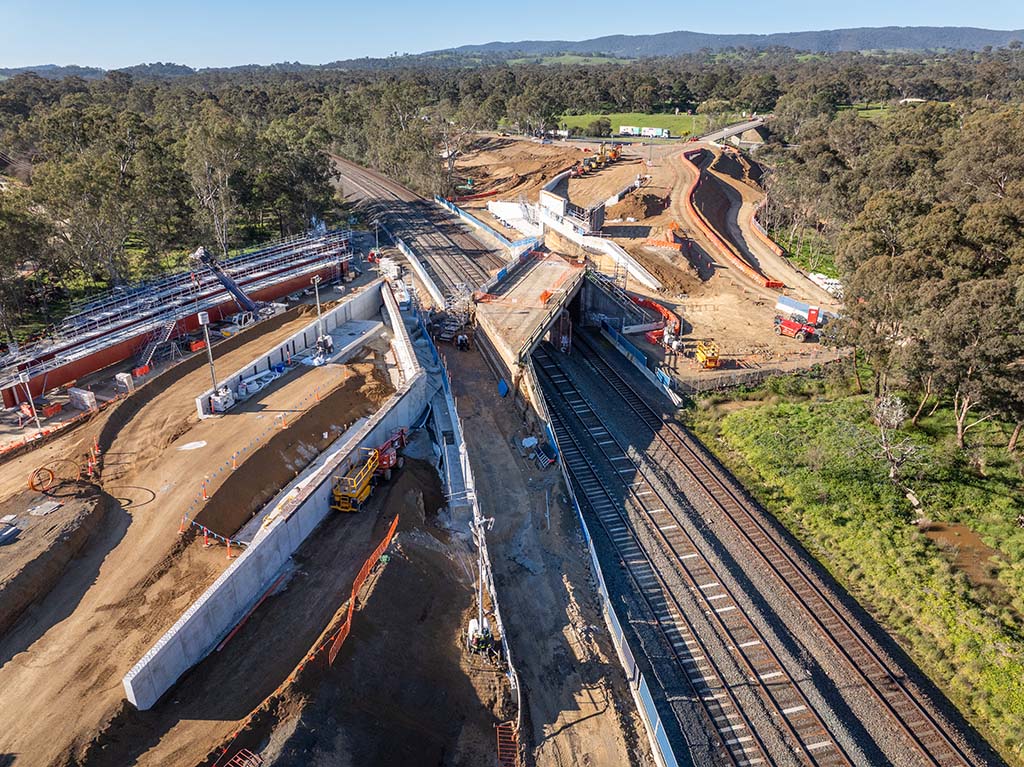 A construction site extends over a railway track which has three tracks. A bridge is being constructed over the tracks. One side of the bridge has been established.