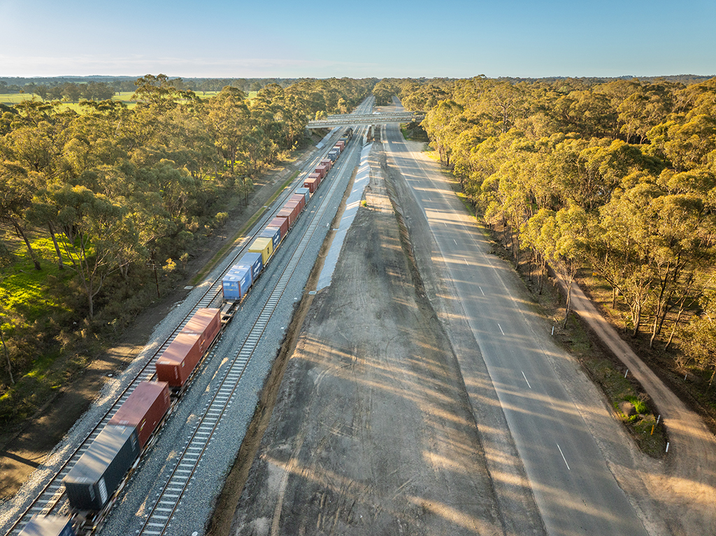 Aerial view of a freight train travelling through a rail corridor beside a road and bushland.
