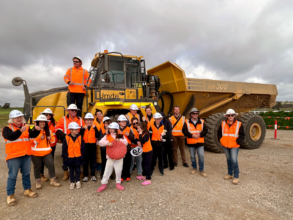 Students and staff wearing high-visibility clothing and hard hats during a site visit beside a large dump truck.