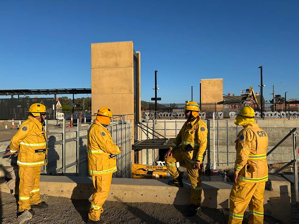 Four people in high visibility protective work suits and helmets stand beside a train station worksite.