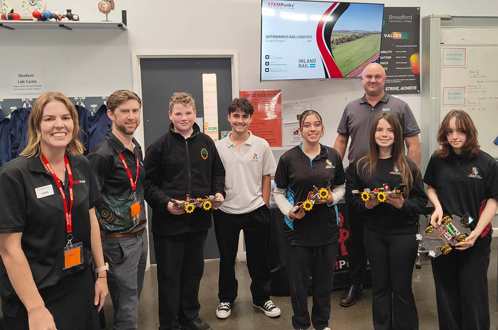 A row of adults and students stand in a classroom. The students are holding small, electronic vehicles.